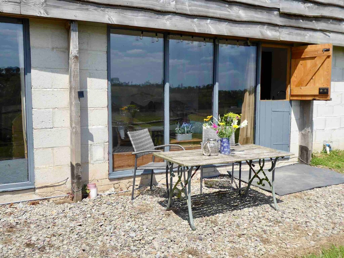 A rustic outdoor table set on gravel is adorned with a vase of fresh flowers, surrounded by a single chair. Large glass doors reflect the natural light from inside, offering a view of the nearby landscape and inviting a connection with the tranquil surroundings.