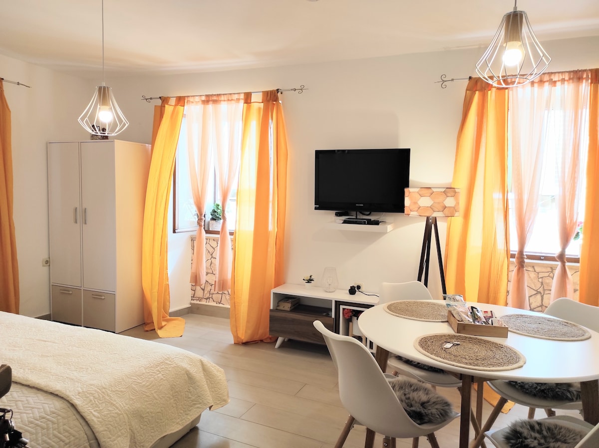 A bright living space is presented with large orange curtains framing two windows, allowing natural light to illuminate the room. A round dining table with white chairs is situated near a modern television mounted on the wall, complemented by stylish pendant lighting above.