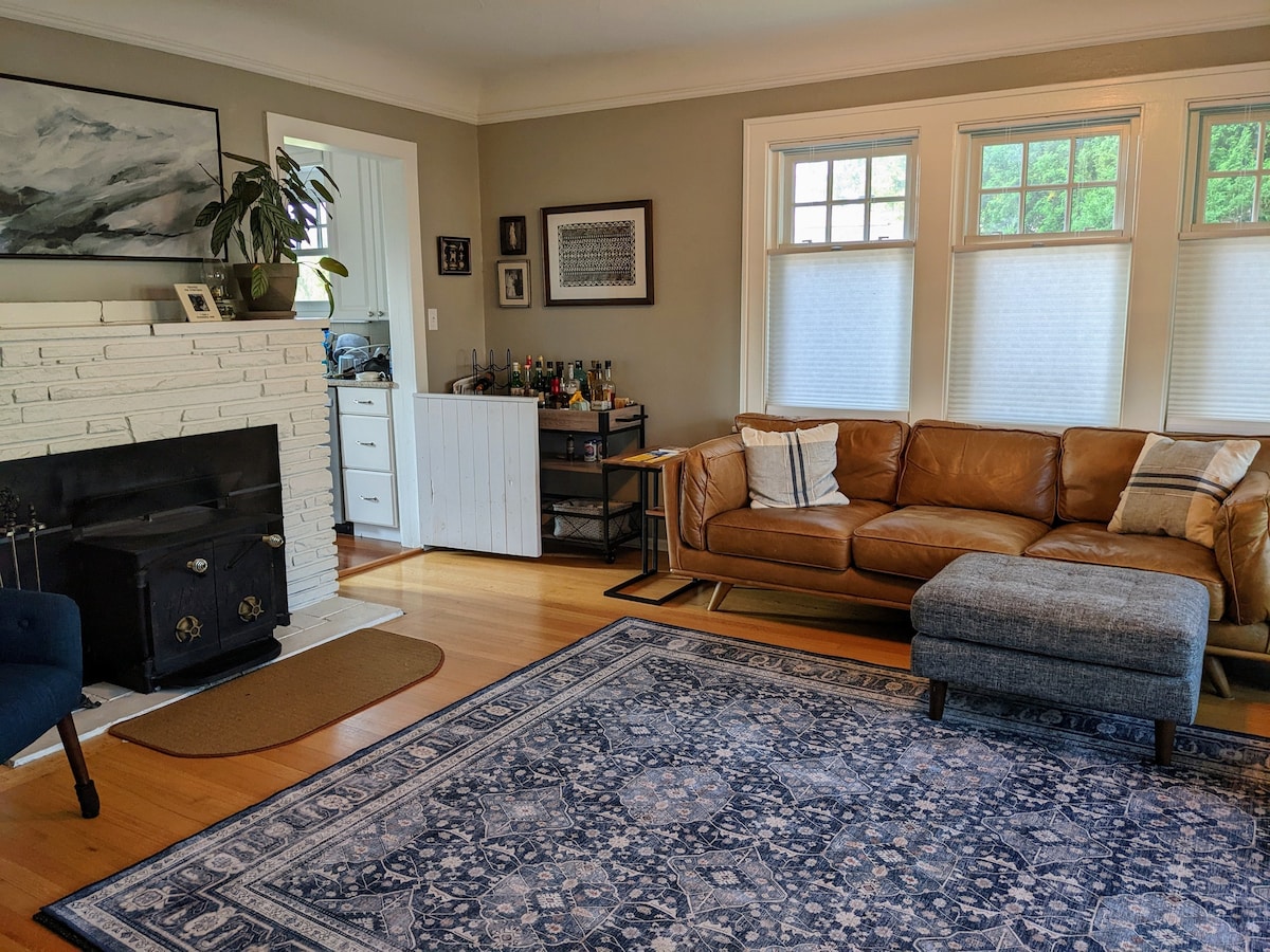 A cozy living room features a brown leather sofa and a matching ottoman, arranged over a decorative area rug. A stone fireplace adds warmth to the space, while large windows allow natural light to brighten the room. A small side table and a plant enhance the inviting setting.