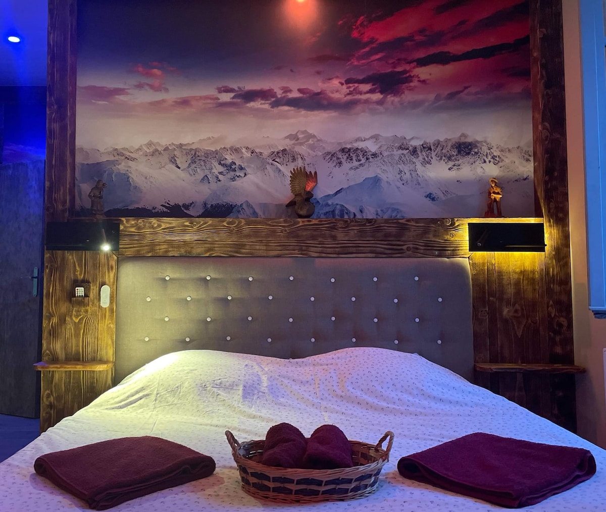 The sleeping area is highlighted by a large, plush bed with a padded headboard. A striking mountain landscape mural adorns the wall above, complemented by warm lighting on either side. Decorative items are placed on wall shelves, adding character to the space.