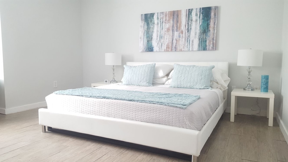 A spacious bedroom features a large bed with a light-colored quilt and decorative pillows. Two bedside tables hold elegant lamps, while a piece of wall art with cool tones adds a serene touch. Natural light enters through a nearby window, enhancing the tranquil atmosphere.