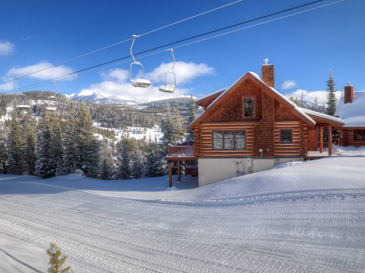 A cozy cabin, characterized by wooden architecture, is set against a snowy landscape. A ski lift is visible in the background, with snow-covered trees surrounding the property. The clear blue sky and distant mountain peaks provide a scenic backdrop.