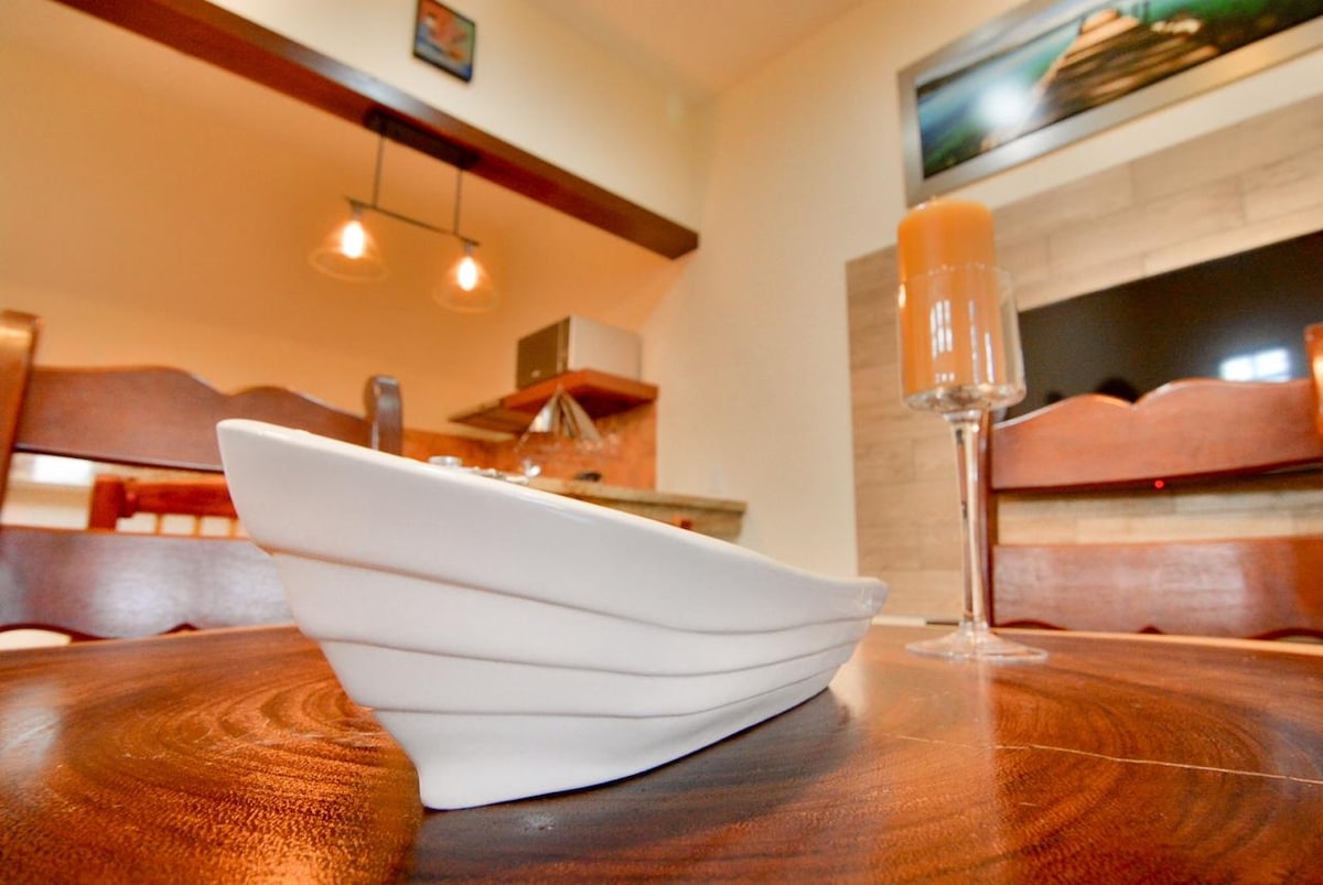 A close-up view of a white ceramic dish rests on a wooden table, complemented by a tall glass filled with a light beverage. The background reveals a softly lit living space, featuring wooden chairs and a wall-mounted television.