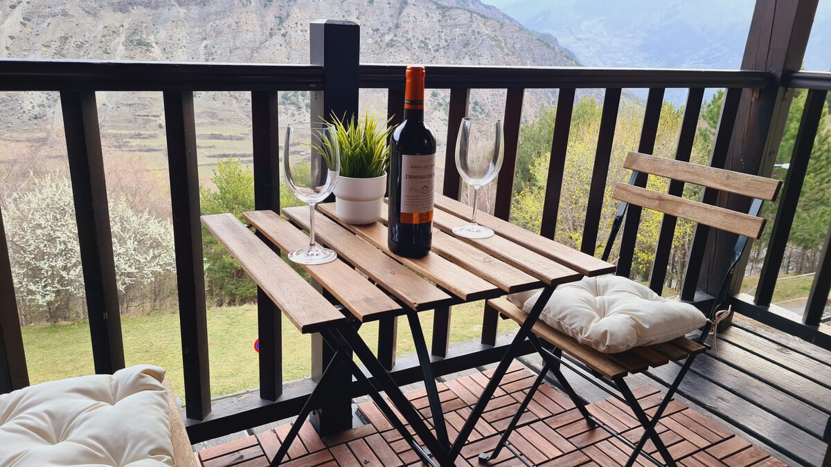 A wooden table is set with two glasses and a bottle of wine, situated on a balcony. Surrounding greenery and a mountainous landscape are visible in the background, enhancing the outdoor dining experience. Cushions add comfort to the seating.