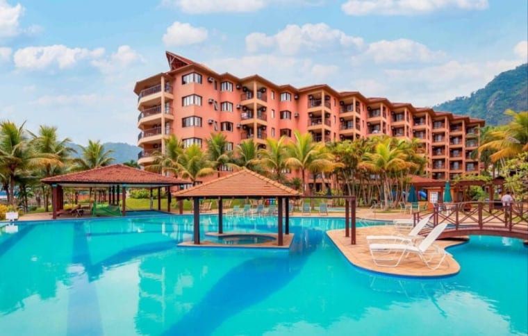 A four-story resort building features a terracotta façade, surrounded by lush palm trees. A large swimming pool with clear blue water is visible in the foreground, alongside wooden sun loungers and shaded seating areas on raised platforms.