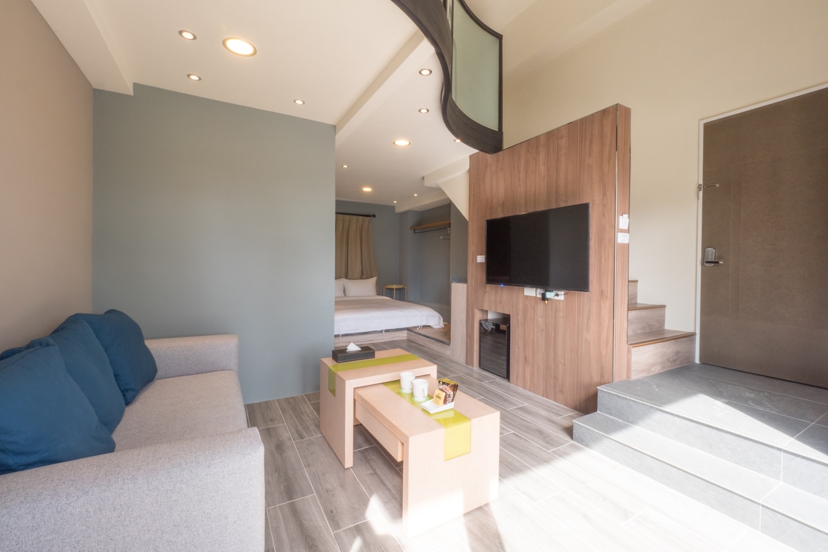 A spacious living area is shown, featuring a light grey sofa with blue cushions and a wooden coffee table. The room is separated by a partial wall with a flat-screen TV mounted. Natural light fills the space, creating a bright and open feel.