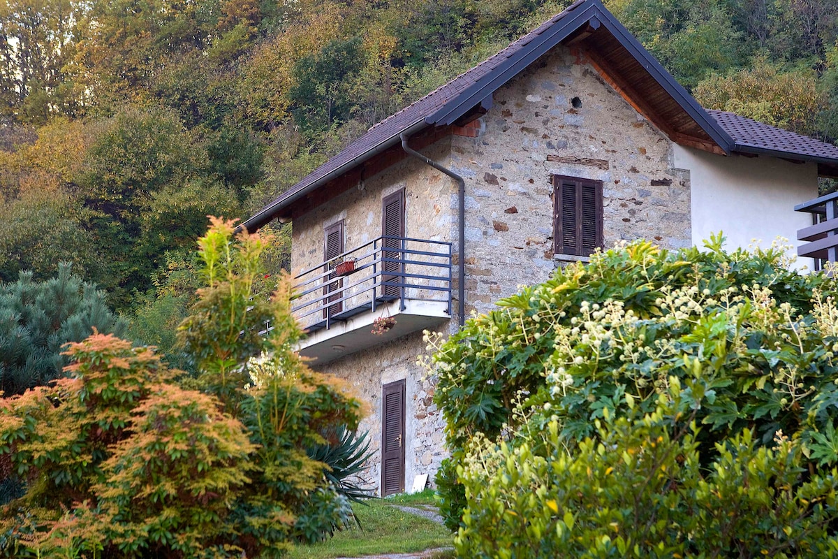 A charming stone house is surrounded by lush greenery, showcasing a warm blend of natural textures and colors. The second floor features a balcony with wooden railings, while large windows invite natural light into the living spaces.