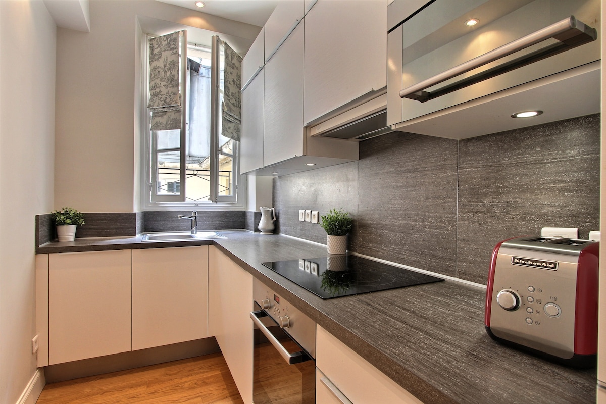 The kitchen features modern cabinetry and ample counter space, with a view from a window opening to the outside. Appliances, including a toaster and a built-in stovetop, are neatly arranged, while small potted plants add a touch of greenery.