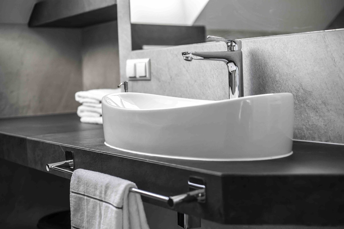 A modern bathroom sink is featured on a sleek countertop, complemented by neatly arranged towels and minimalist fixtures. The bowl-shaped sink is surrounded by smooth surfaces, emphasizing a clean and functional design. Soft lighting accentuates the textures and lines of the space.