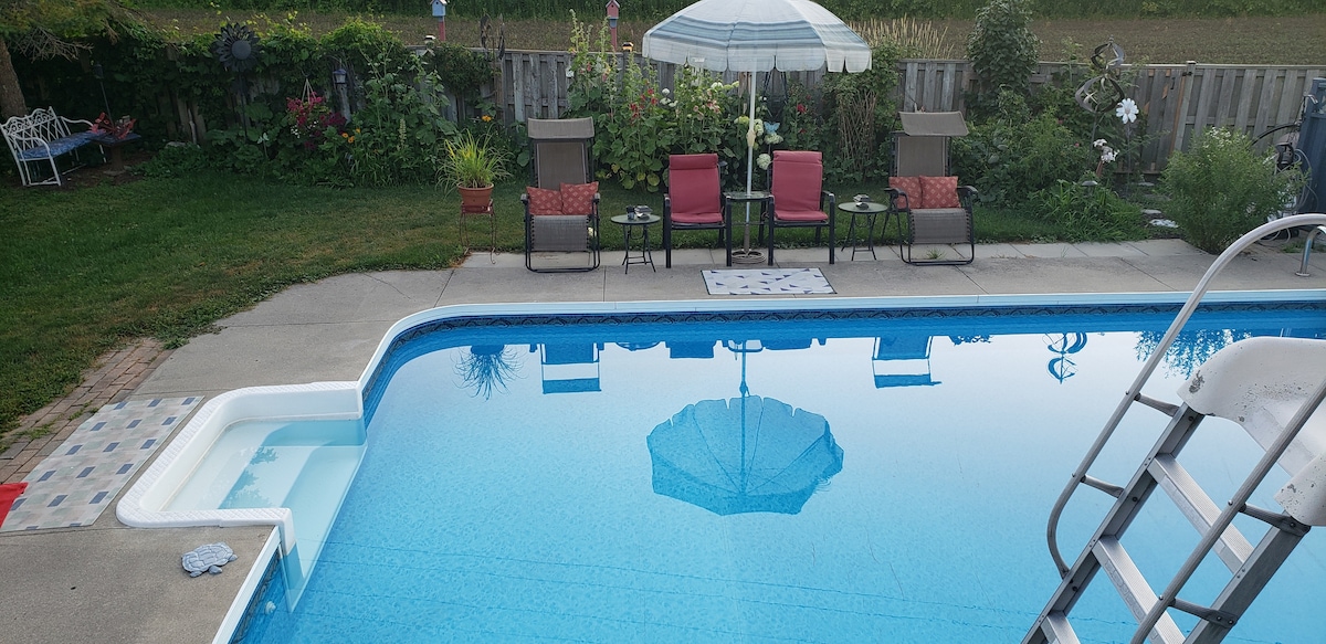 The seasonal inground swimming pool is surrounded by a well-maintained garden. Lounge chairs and small tables are positioned under a large umbrella, providing shaded seating. The pool features a shallow entry and clear blue water, reflecting the surrounding greenery.
