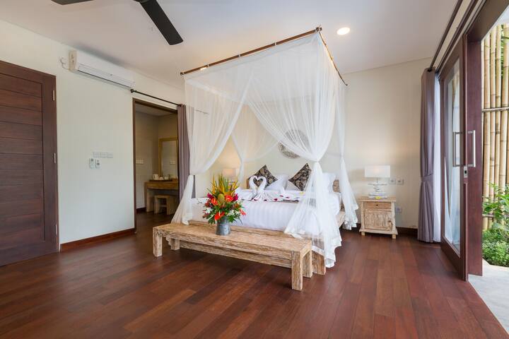 Airy bedroom with a four-poster bed draped in sheer white curtains, creating a dreamy and romantic space.  The room features hardwood floors and opens onto a balcony with scenic views.