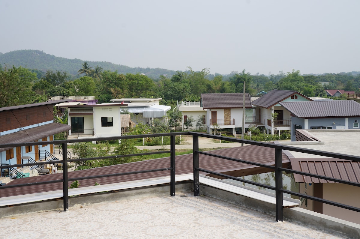 A rooftop terrace offers views of neighboring houses nestled among greenery, with distant hills visible on the horizon. A railing surrounds the terrace, providing a boundary while showcasing the expansive outdoor area.