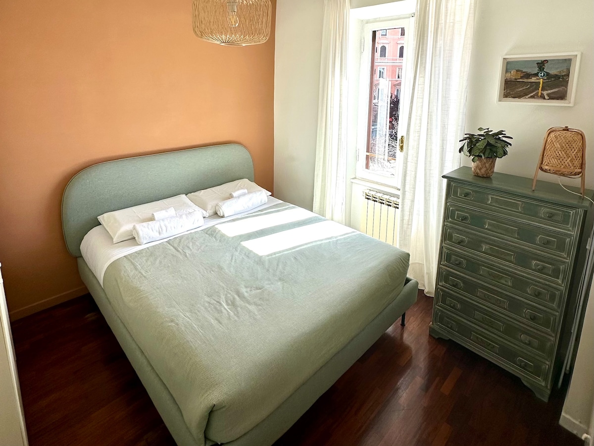 A well-lit bedroom features a spacious bed with soft green upholstery and neatly arranged pillows. A large window dressed in light curtains allows natural light to brighten the room. A tall, vintage dresser and a small plant add a touch of style to the serene space.