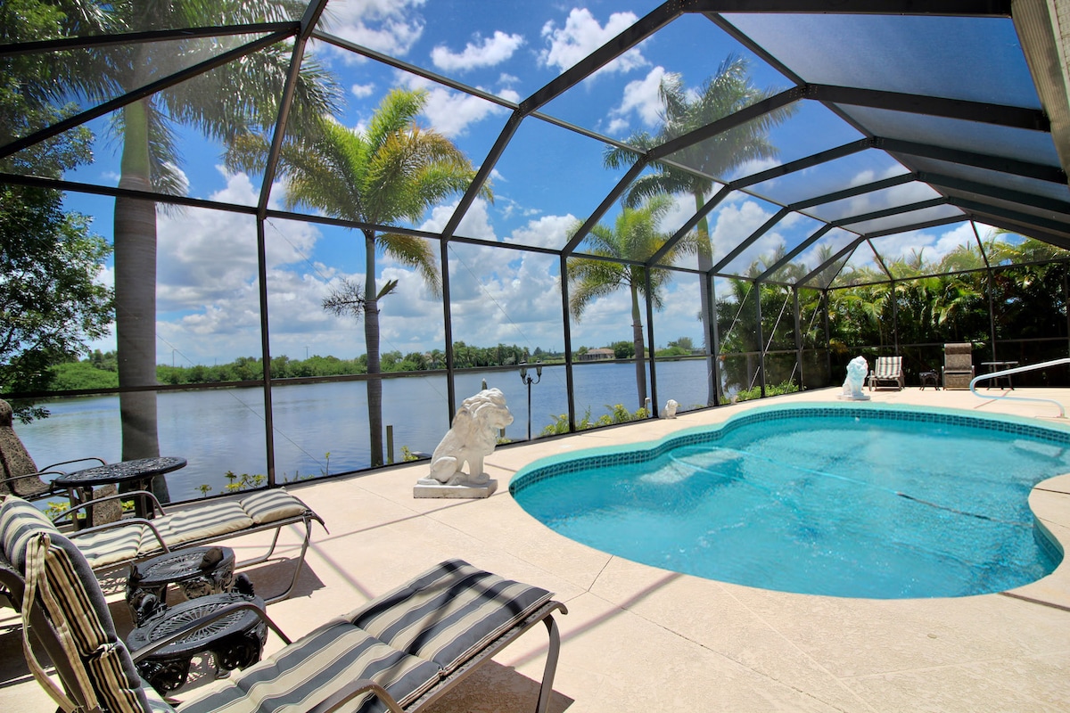 The pool area is enclosed by a mesh screen, offering views of the tranquil Valmora Lake. Sun loungers are positioned around the pool's edge, complemented by palm trees and tropical plants in the background under a clear blue sky.