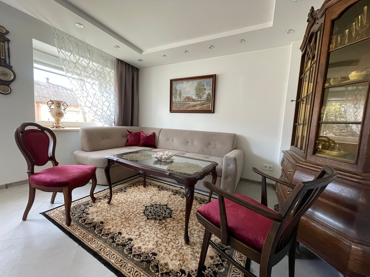 A well-appointed living area features a light-colored sectional sofa and an elegant wooden coffee table. Vintage-style chairs with red upholstery surround the table. Natural light filters through sheer curtains, enhancing the space. A decorative painting adorns the wall, contributing to the room's refined ambiance.