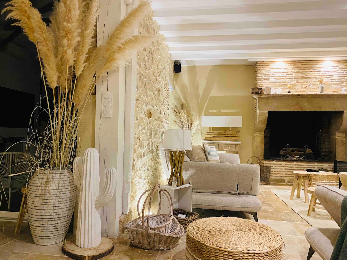 A welcoming living area features textured stone walls and wooden beams overhead. A large fireplace acts as a focal point, surrounded by comfortable seating. Natural elements like pampas grass and a cactus sculpture add a touch of warmth, while woven baskets and a round coffee table complete the serene ambiance.
