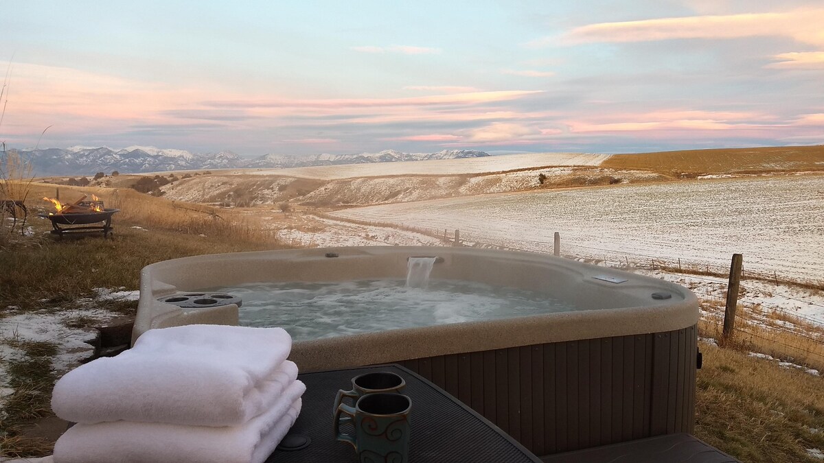 A private hot tub sits on a patio, surrounded by scenic views of rolling hills and distant mountains. Towels are neatly stacked nearby, and a steaming mug rests on the table, suggesting a tranquil experience against a backdrop of a pastel sunset sky.