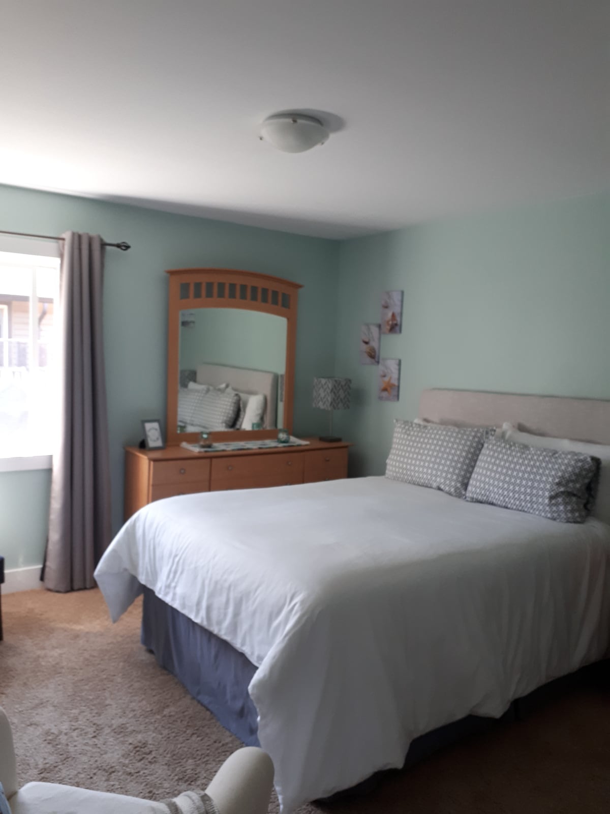 A bright guest room is presented with soft green walls and natural light filtering in through a window. A spacious bed with cozy bedding rests against the wall, complemented by a wooden dresser featuring a mirror. Decorative pieces add a subtle charm to the room.