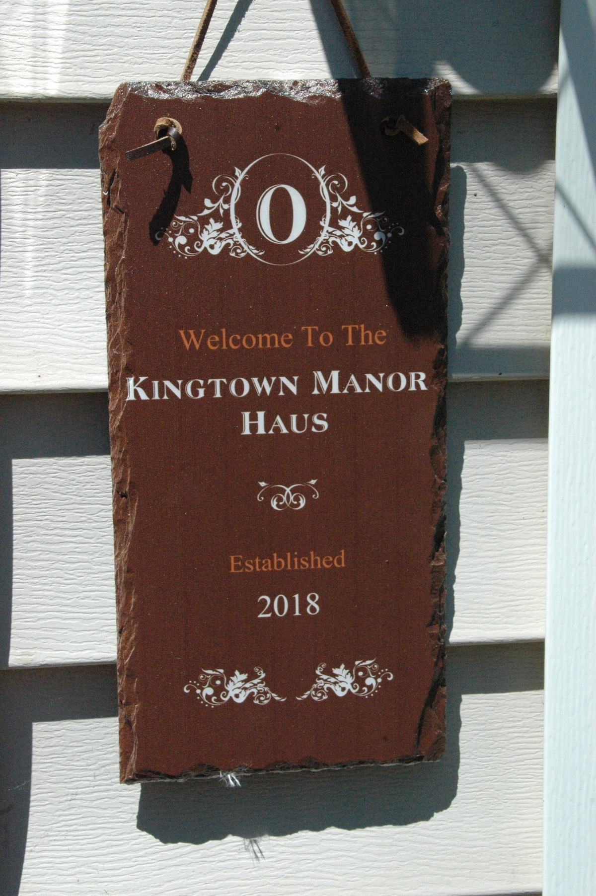 A welcome sign is mounted on the exterior wall, featuring 'Welcome To The KINGTOWN MANOR HAUS' prominently displayed. The sign is crafted from slate, with a rustic look and the year 'Established 2018' included at the bottom.