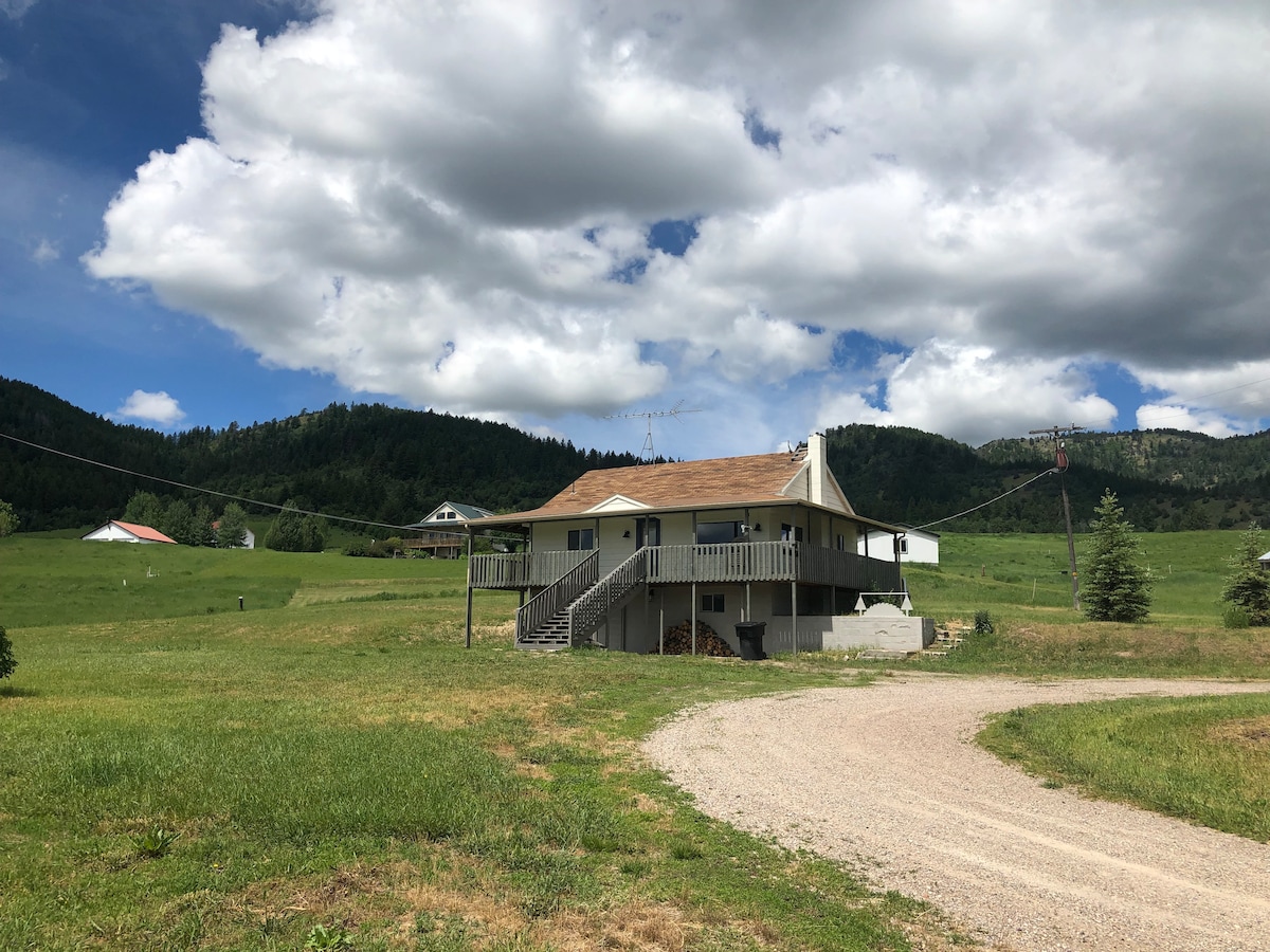 The farmhouse is situated on a spacious grassy area, surrounded by rolling hills and trees. A gravel driveway leads to the front steps, and a large deck is visible on the upper level. The sky is partly cloudy, enhancing the rural scenery.