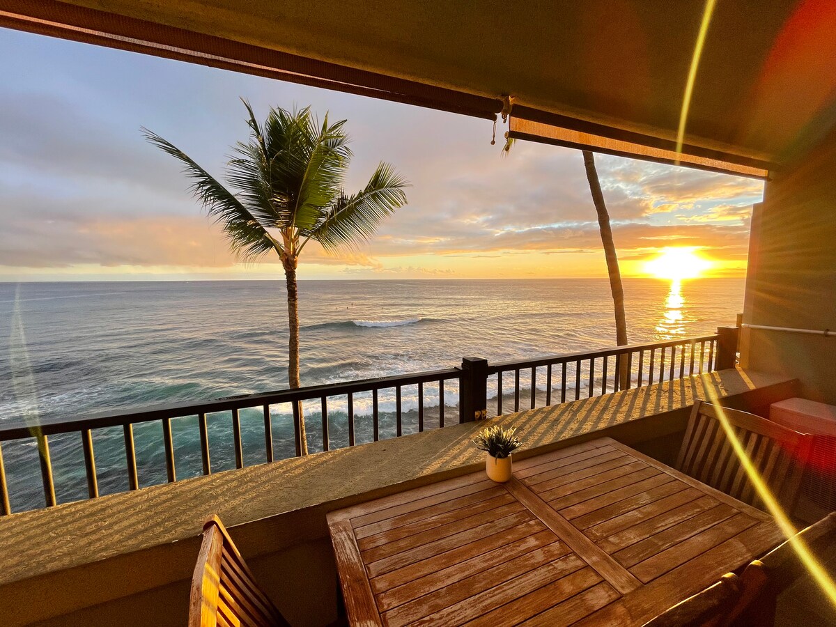 An outdoor lanai is shown with a wooden dining table and chairs. A palm tree is visible beside the ocean, where the sun is setting over calm waters. The railing provides an unobstructed view of the horizon and the colorful sky.