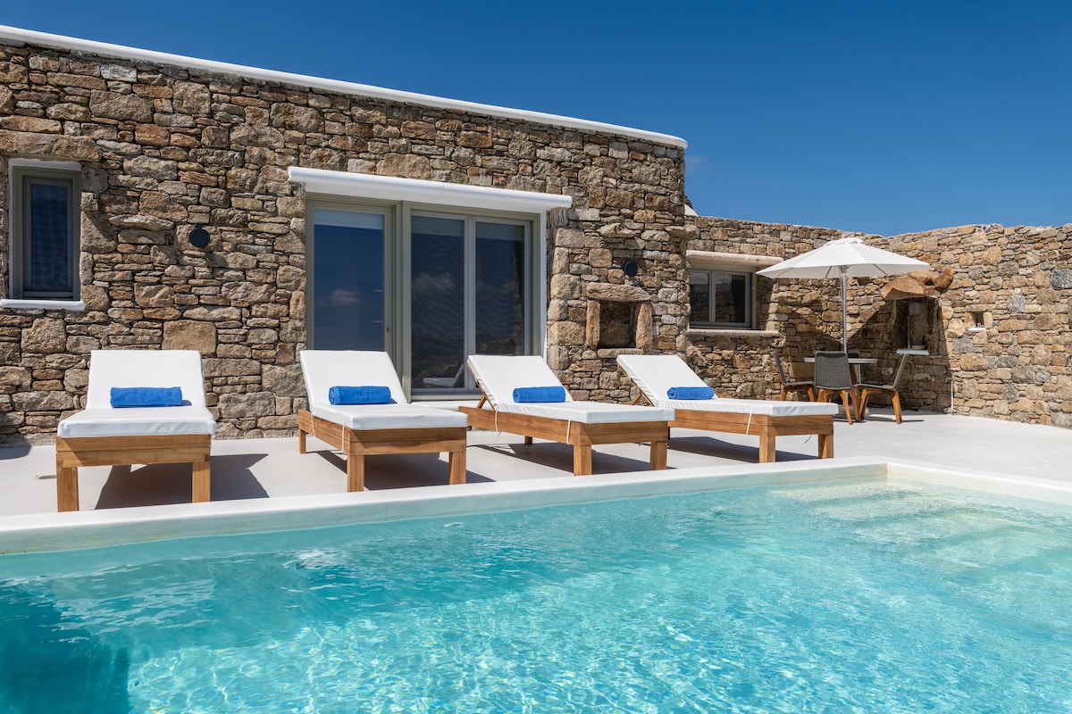 A private terrace showcases a tranquil infinity pool with clear waters. Sun loungers with blue towels are arranged around the pool. A sun umbrella and a small dining table are positioned nearby, set against the backdrop of traditional stone architecture.