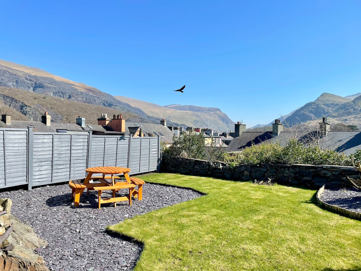 A private garden features a picnic table set on a grassy lawn surrounded by stone borders. The view includes rolling hills and mountains under a clear blue sky, while a bird soars in the distance, adding to the serene atmosphere.
