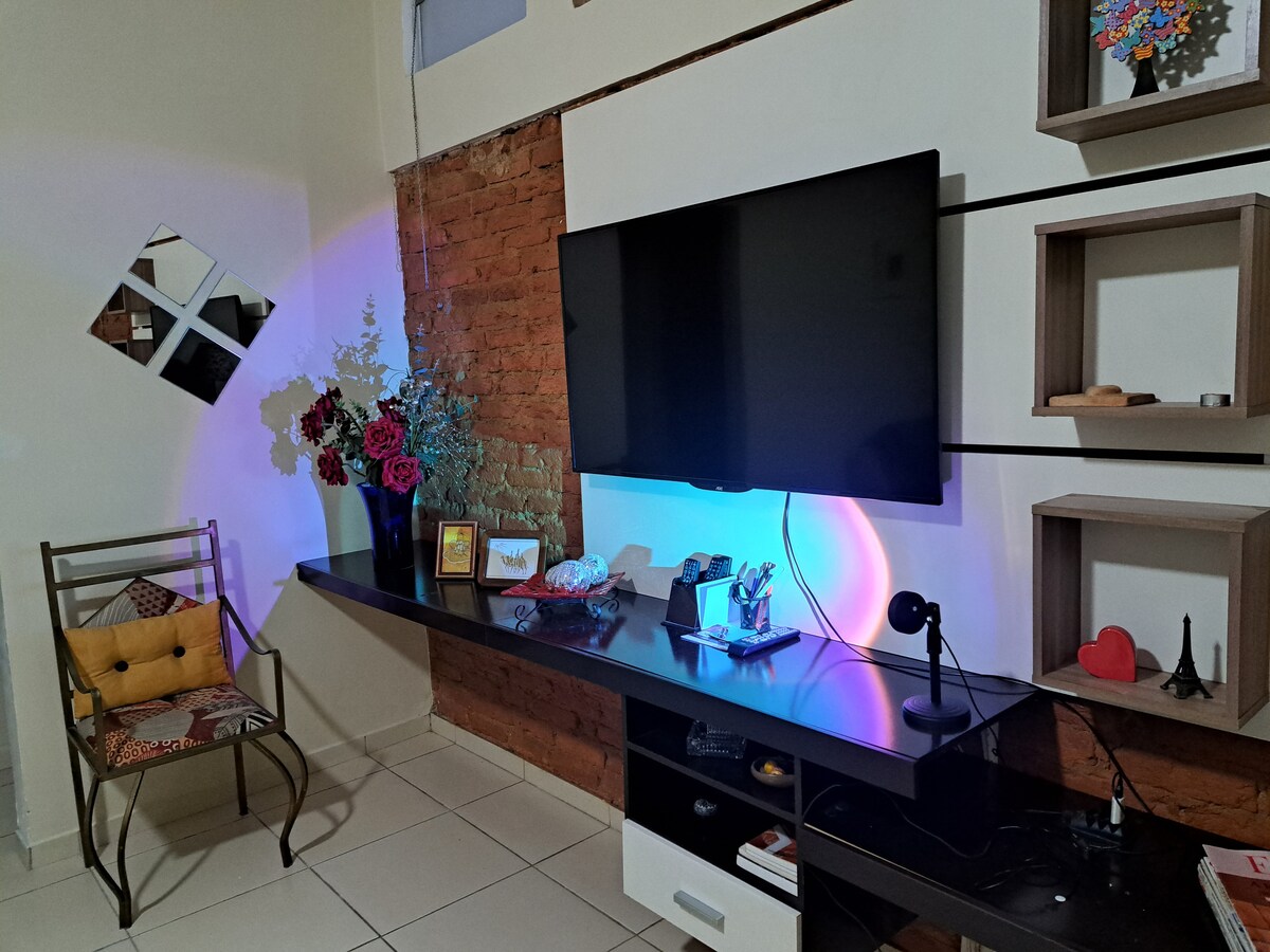 A welcoming living area features a modern wall-mounted TV above a sleek black shelf. Decorative elements include a small chair with a colorful cushion and various adorned shelves holding an assortment of items. Soft lighting creates an inviting ambiance against the exposed brick wall.