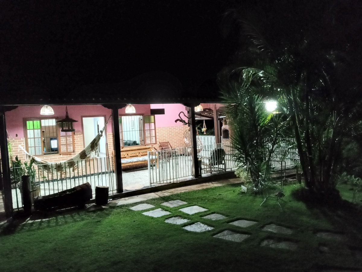 A nighttime view of a rural retreat showcases a cozy porch illuminated by soft lighting. Seating areas are visible, including a hammock and a wooden sofa set. Lush green plants surround the space, while stepping stones lead through the grassy area.
