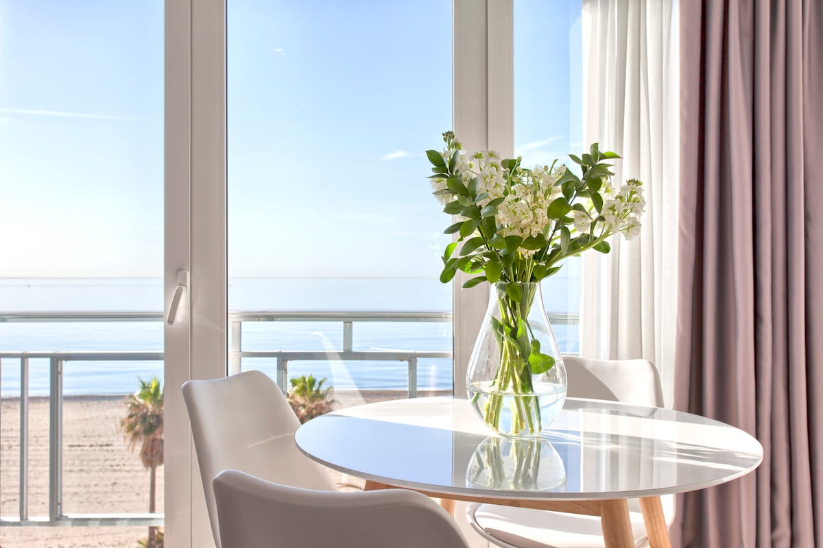 A round glass table with a wooden base is set near large windows offering unobstructed sea views. A vase filled with fresh white flowers sits at the center, complemented by white chairs. Sheer curtains frame the scene, allowing natural light to brighten the space.