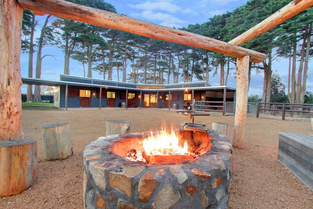 A stone fire pit is prominently positioned in an open outdoor area, surrounded by wooden seating. Tall trees frame the space, providing a natural backdrop to the cozy gathering spot, with the main building visible in the distance, inviting relaxation and outdoor dining.