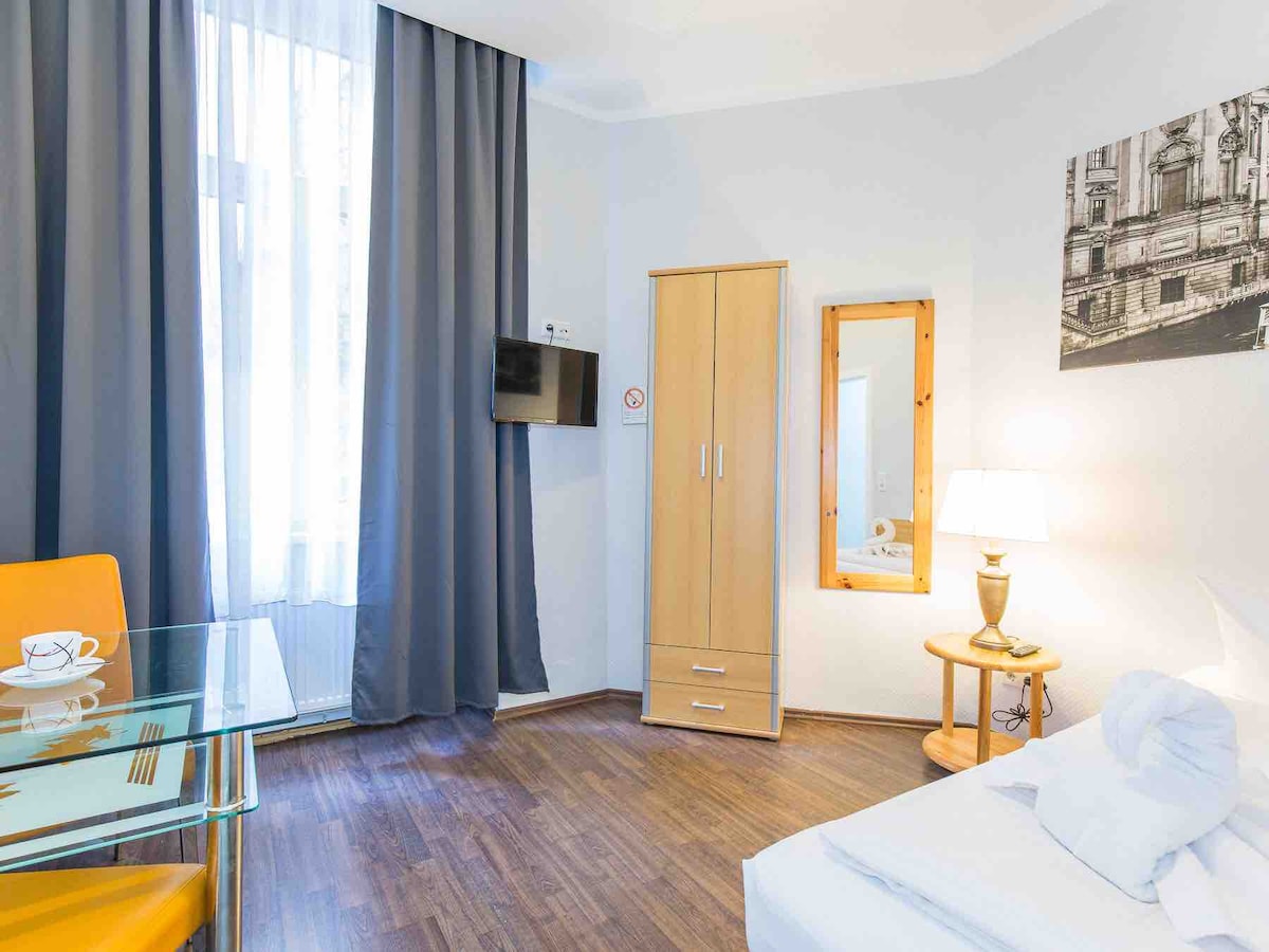 A modern room features a spacious bed complemented by a bedside lamp. A tall wooden wardrobe and a wall-mounted TV are present. A glass dining table with a yellow chair and a mirror are visible, alongside large windows draped with dark curtains, allowing natural light to enter.