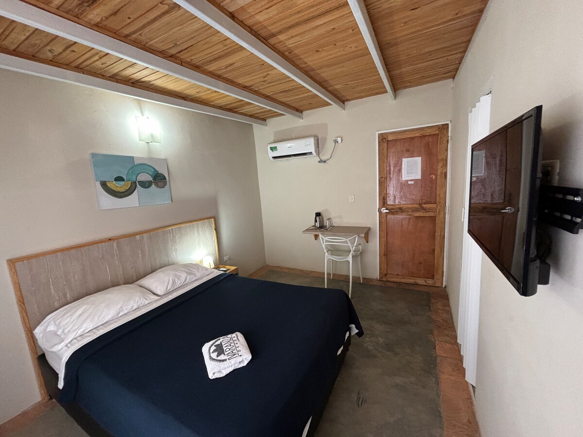 A well-maintained guest room features a spacious bed with a navy blue coverlet. Natural wood accents are visible in the ceiling and furnishings. A small desk with a kettle is positioned near the door, while a flat-screen TV is mounted on the wall, providing modern amenities.