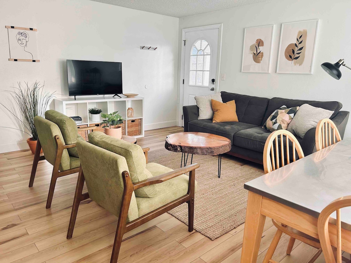 A bright living area features a dark sectional sofa adorned with decorative pillows, accompanied by two green accent chairs. A wooden coffee table sits in the center, and a television is mounted on the wall opposite. Natural light enters through the door, enhancing the cozy ambiance.