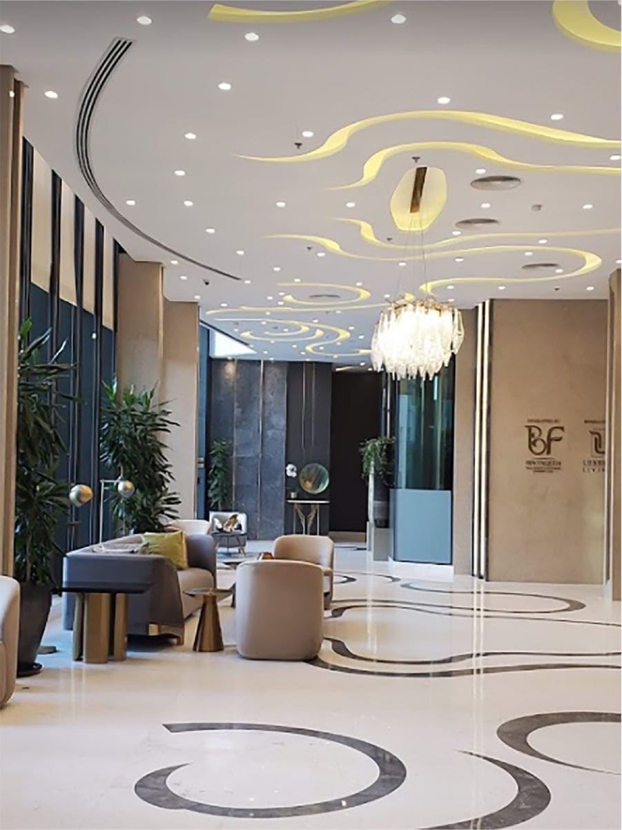 A modern lobby features a sleek design with elegant lighting and a large chandelier. Plush seating arrangements are positioned near tall windows, allowing natural light to fill the space. Decorative plants add a touch of greenery to the sophisticated atmosphere.