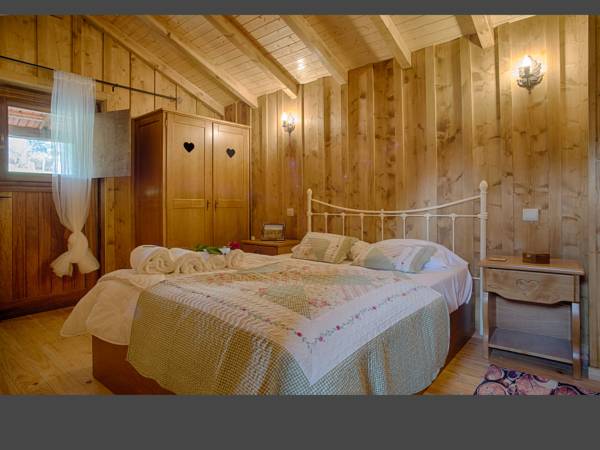 A cozy bedroom features wooden walls and a sloped ceiling, enhancing a natural ambiance. A spacious bed is dressed with a patterned quilt and accompanied by soft pillows. A bedside table holds a small lamp, and a window offers natural light with light drapes.