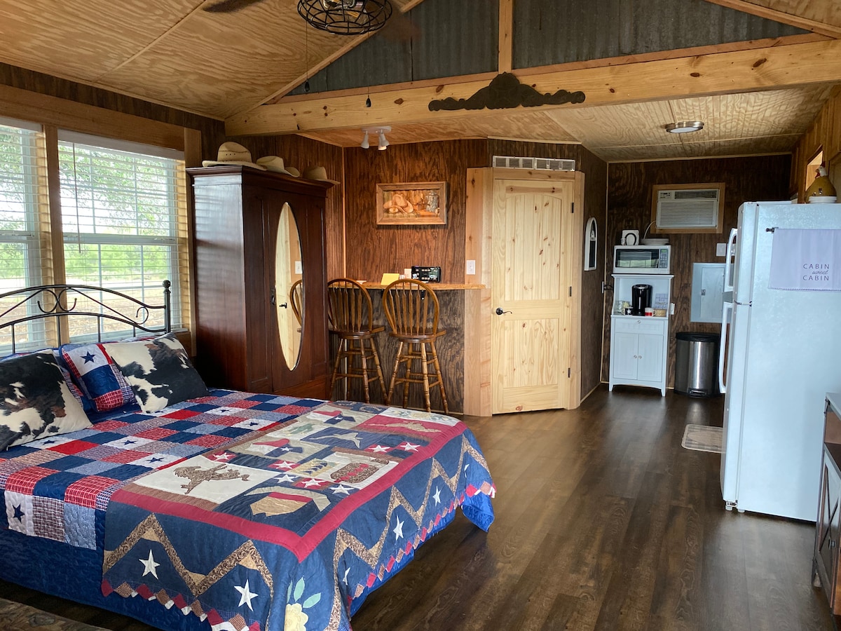 A cozy sleeping area features a bed adorned with a patchwork quilt, incorporating shades of blue and red. Natural light fills the space through large windows, and a small kitchen area is visible, equipped with a refrigerator and coffee station. Wooden accents add warmth to the environment.