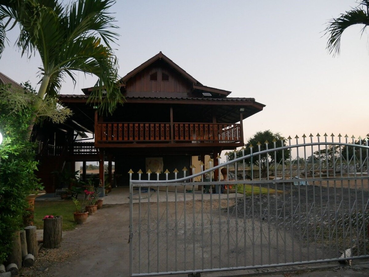 A wooden house with a steep roof is presented, featuring a spacious balcony adorned with railing. A gated entrance leads to a gravel driveway lined with side planters. Surrounding vegetation is visible, complemented by the soft evening light.