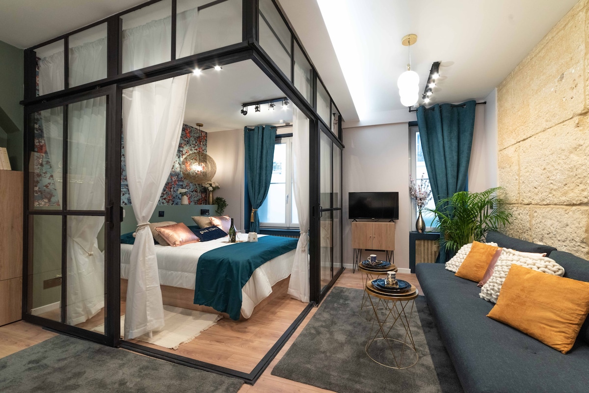 A spacious and stylish room is highlighted by a partitioned sleeping area featuring a large bed covered with a teal duvet. The space includes a comfortable sofa, a coffee table, and an entertainment unit, with natural light streaming through sheer curtains and decorative blue curtains.