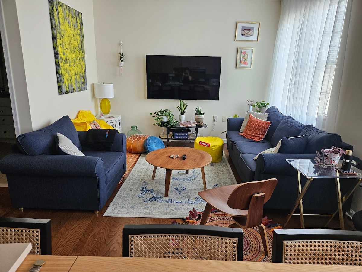 A colorful living area features a dark blue sectional sofa and a wooden coffee table positioned on a decorative rug. Bean bags in bright colors are scattered throughout, alongside a flat-screen TV mounted on the wall. Natural light filters through sheer curtains, enhancing the inviting atmosphere.