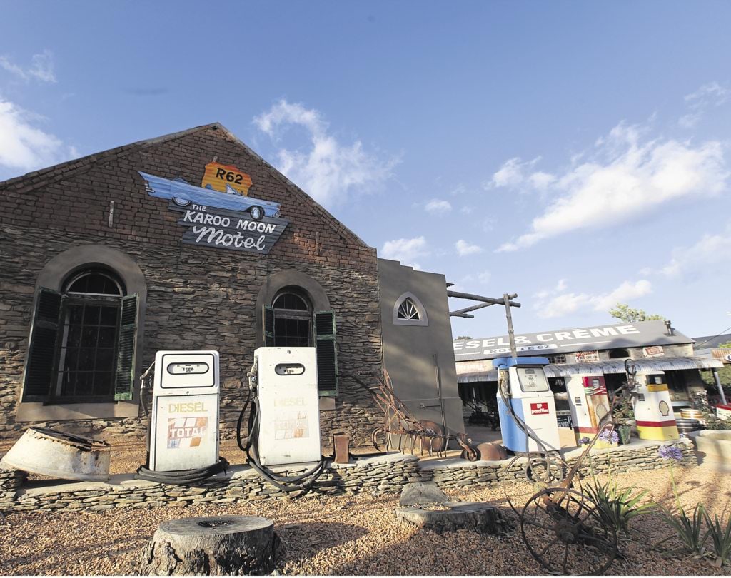 The historic facade of The Karoo Moon Motel displays rustic stonework, accentuated by green shutters and a vintage sign. The building is complemented by a small outdoor dining area, while retro fuel pumps add character to the surrounding landscape.