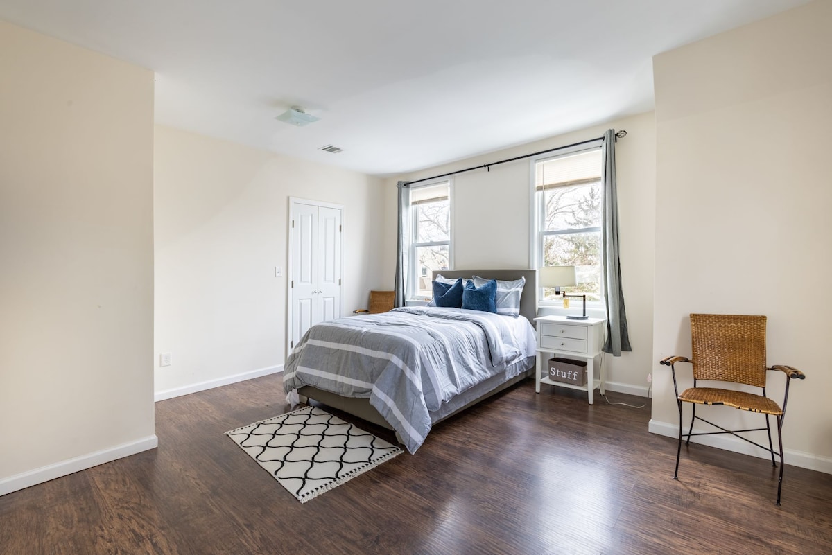 A spacious bedroom features a queen-size bed with a striped blanket and supportive pillows. Two windows bring in natural light, illuminating the room. A small nightstand and a chair are present, along with a patterned area rug on the wood floor, providing a calm and inviting ambiance.
