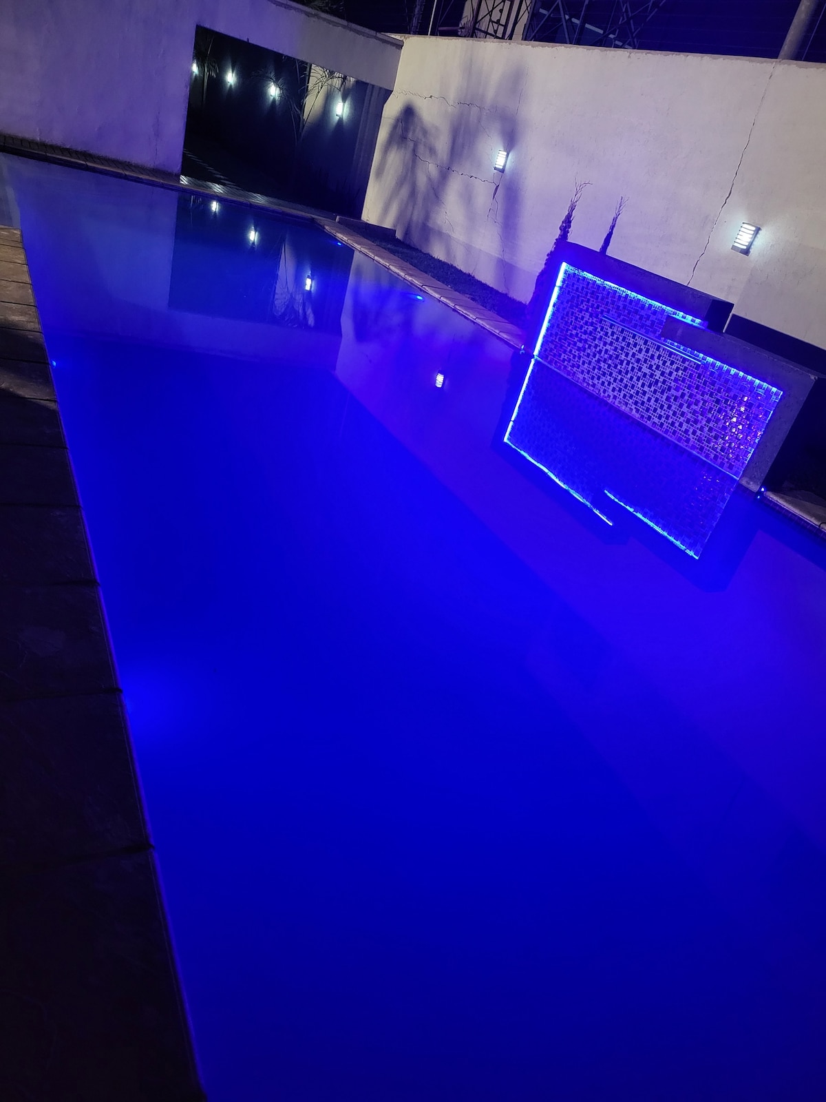 A serene pool area is illuminated by soft blue lights, reflecting on still water. Modern lighting fixtures are mounted on a nearby wall, adding to the tranquil ambiance. The clean lines of the pool create a calming focal point in the space.
