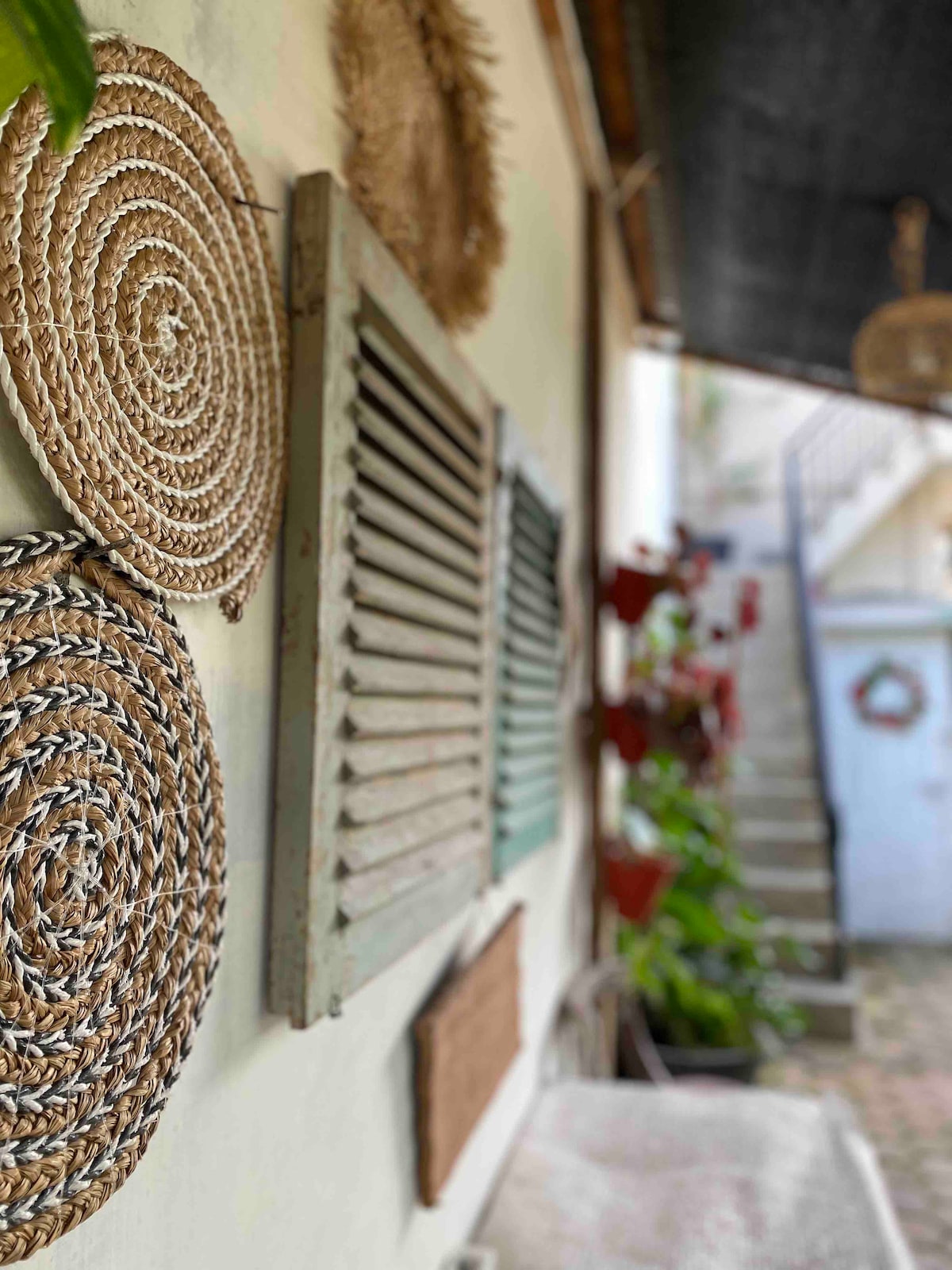 Natural woven decor appears on the wall, accompanied by a set of wooden shutters. In the background, a staircase is visible, leading to the upper level, adorned with greenery and decorative elements, contributing to a welcoming aesthetic.