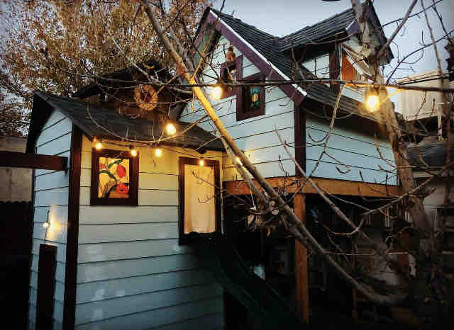 The exterior of a charming blue casita is highlighted by string lights and a unique stained glass window. An inviting slide is visible beside the entrance, framed by a tree with bare branches, creating a whimsical atmosphere.