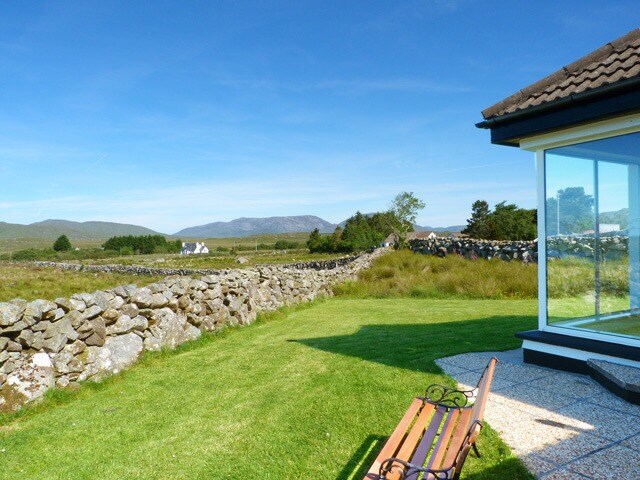 A view of the well-maintained garden area showcasing a wooden bench. Beyond, dry stone walls separate the landscape, which features rolling hills and distant mountains under a clear blue sky.