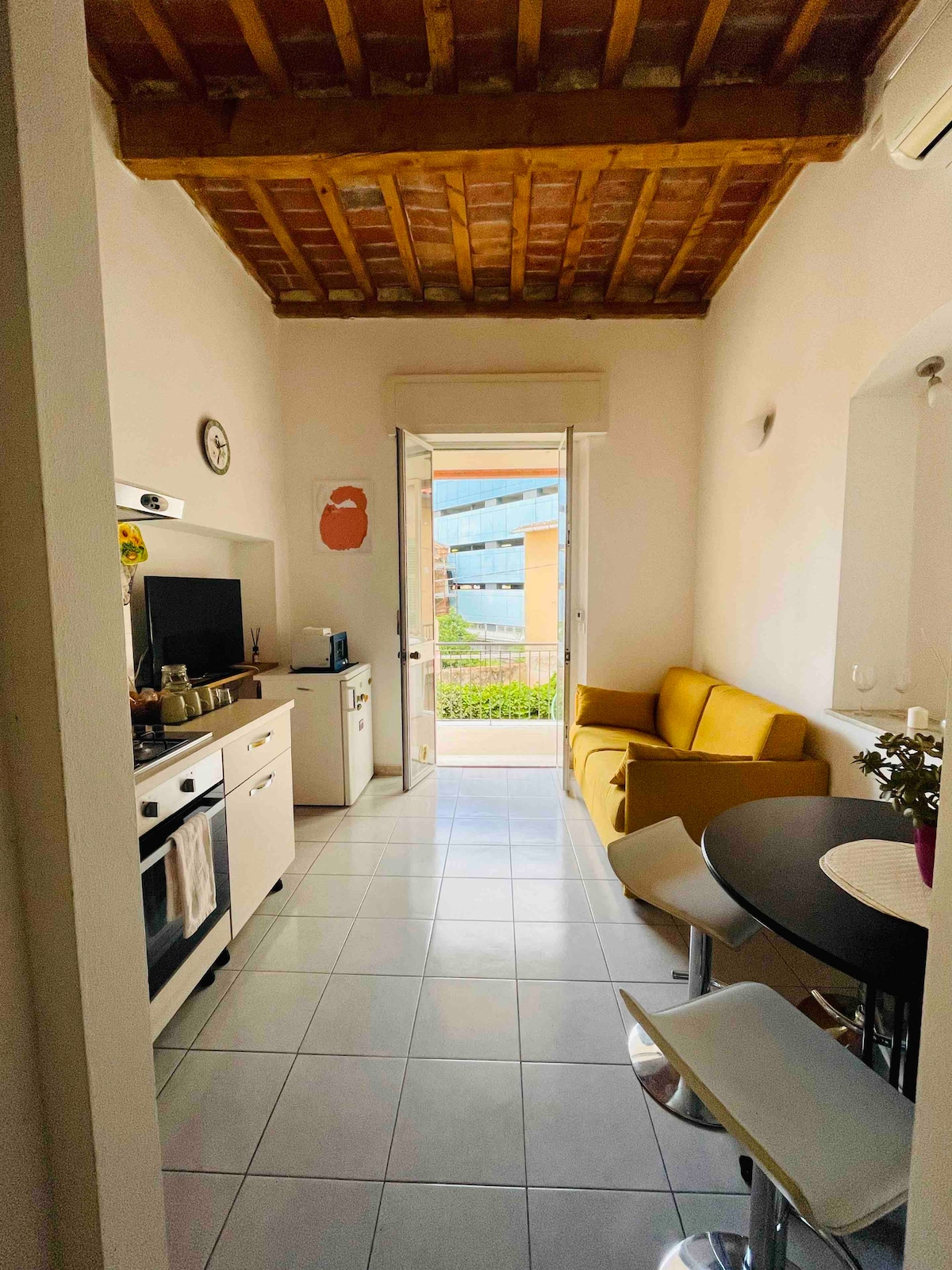 A bright kitchenette leads to a small balcony, with a view of a vibrant outdoor space. A yellow sofa complements the modern black dining table and white bar stools. Natural light bathes the room, enhanced by a wooden beam ceiling.