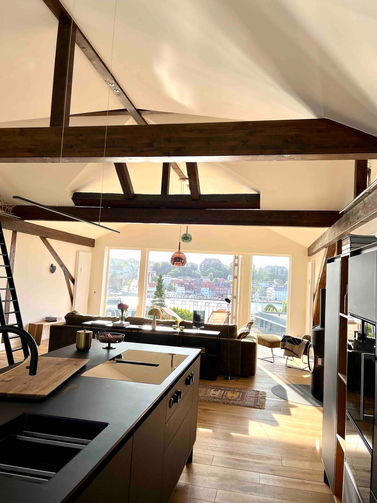 A spacious kitchen is shown, featuring a modern black countertop and open layout. Large windows provide extensive views of the harbor and city. Wooden beams add a rustic element, while comfortable seating is arranged nearby, inviting relaxation and enjoyment of the scenery.