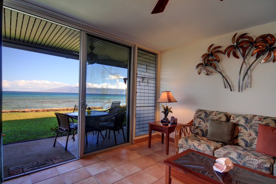 The sitting area features a comfortable sofa adorned with tropical-patterned cushions. A wooden coffee table sits in front, alongside a lamp providing soft light. Floor-to-ceiling sliding glass doors open to a lanai with ocean views, seamlessly connecting indoor and outdoor spaces.
