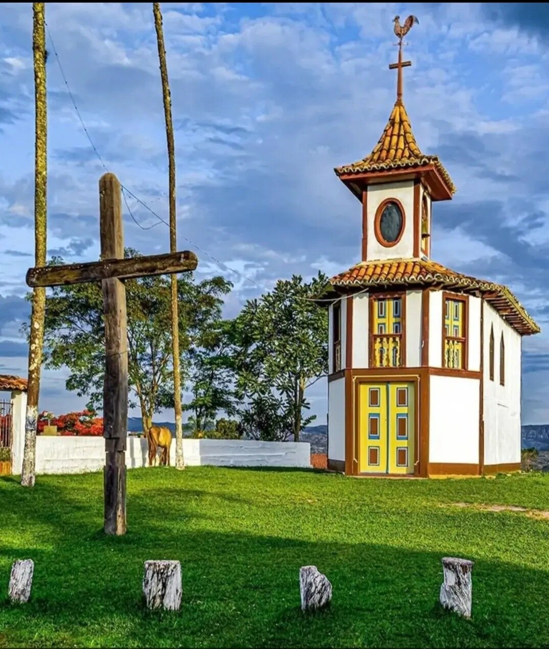 A charming white structure with a brown and yellow facade features a pointed rooftop and a round window. The building is accompanied by a large wooden cross and lush green grass, with trees and a cloudy sky visible in the background.